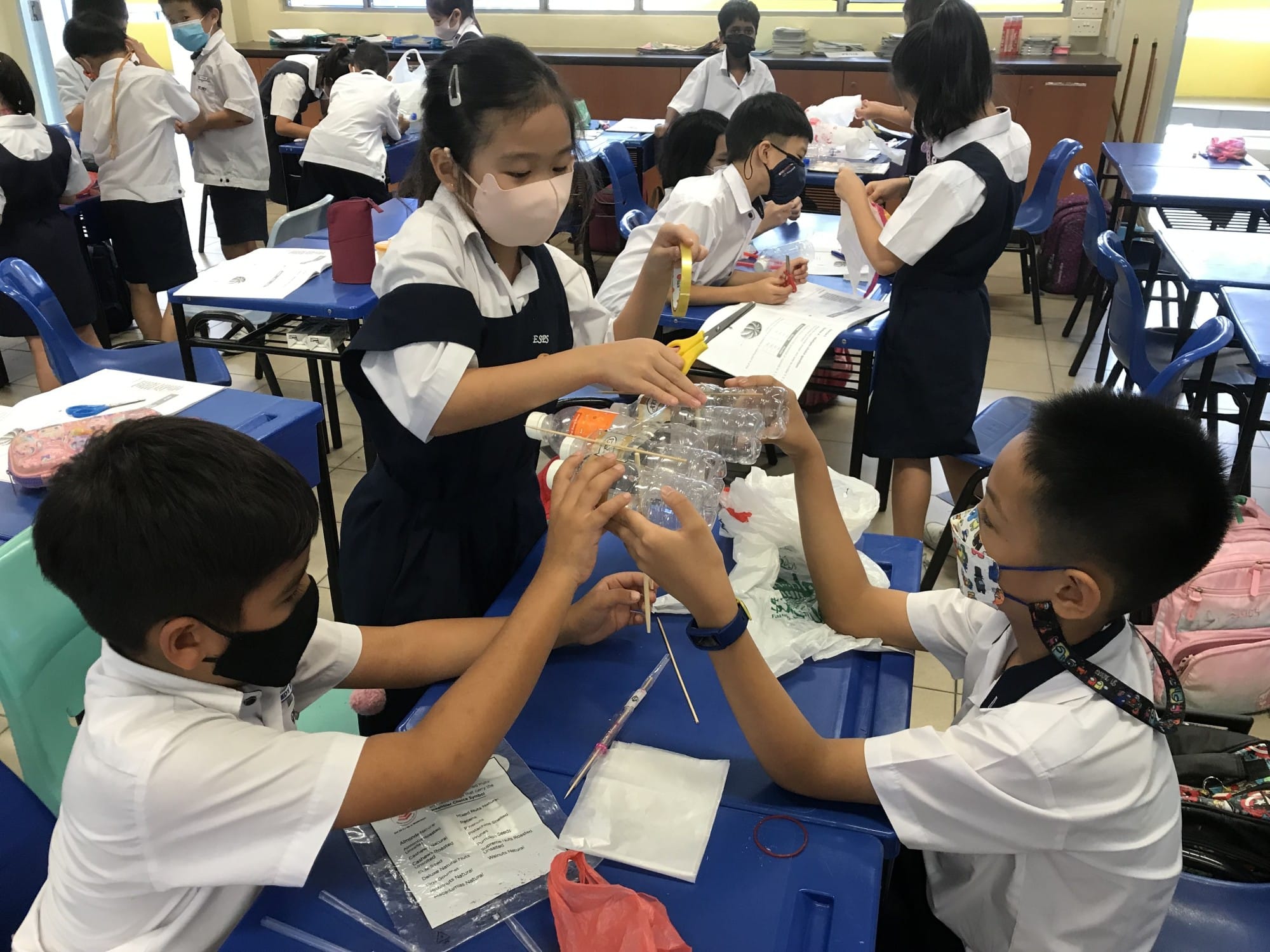 Three masked students in school uniforms build a structure using plastic bottles and sticks at a blue desk in a classroom.